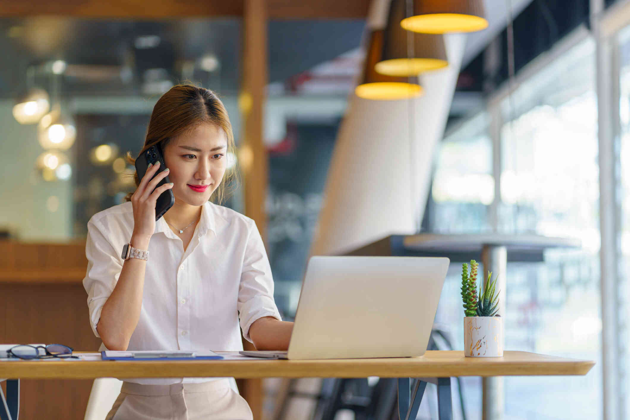 A woman in a button down shirt sits at a table with her laptop open infront of her as she talks on the phone.