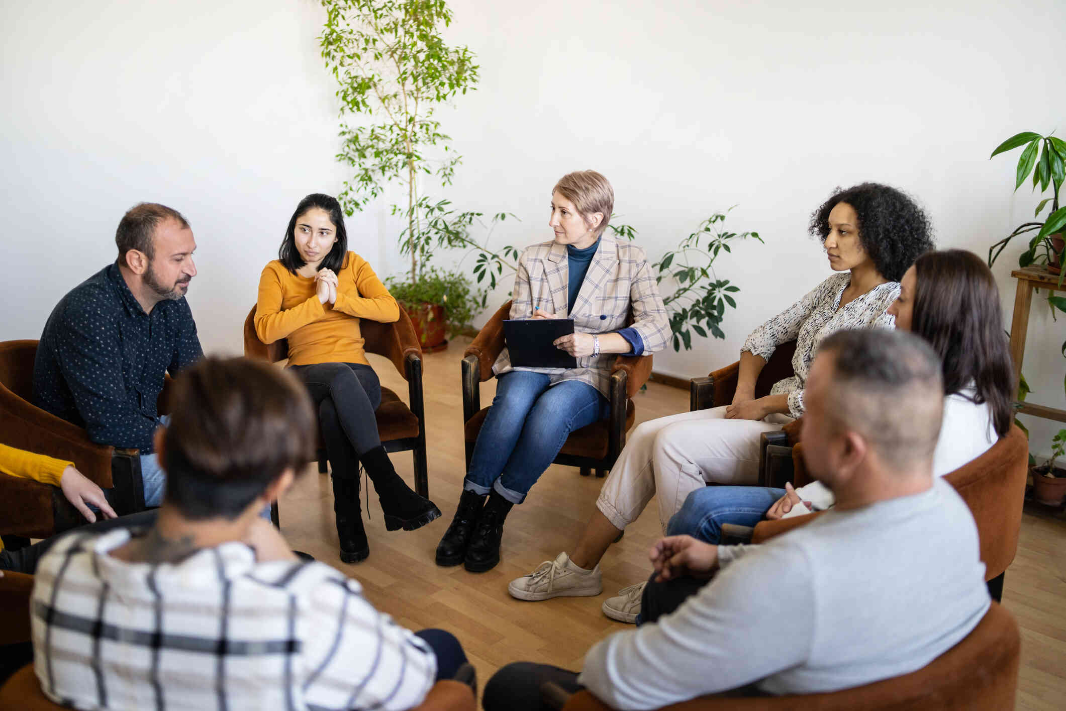 A group of a dults sit in chairs in a circle with a female therapist and talk during a group therapy session.