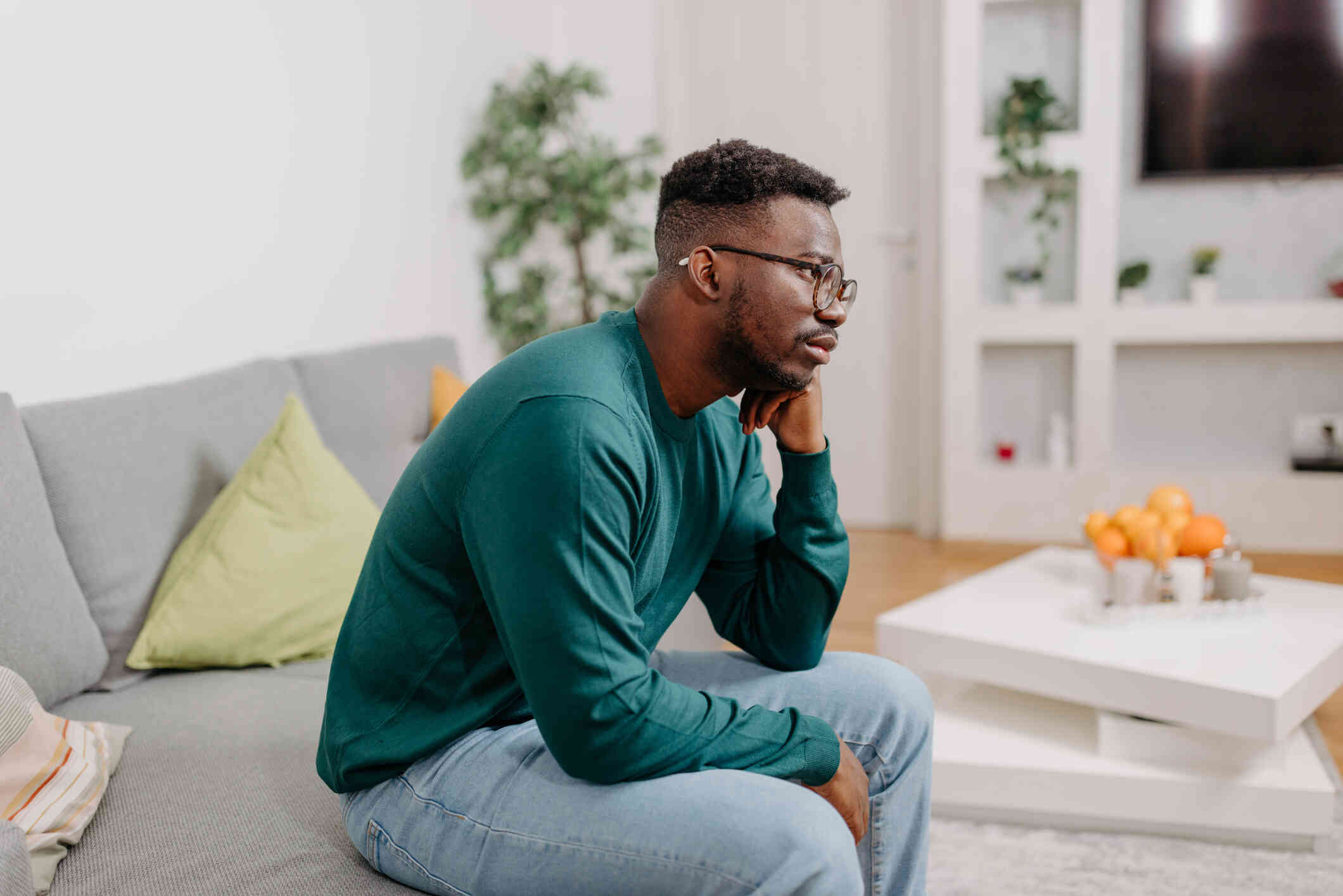 A man in a green shirt sits slouched over on the couch with his head resting in his hand as he gazes off sadly.