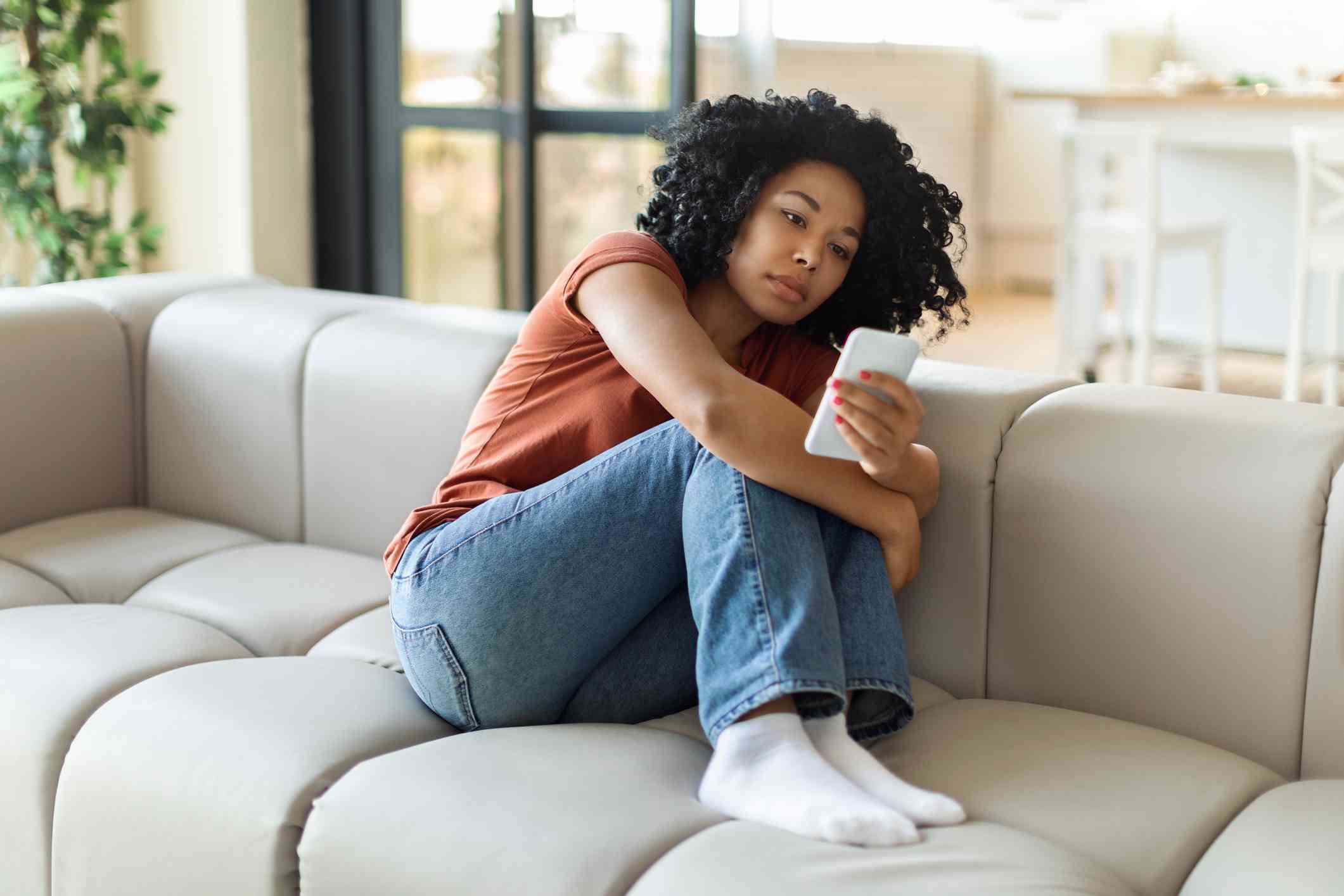 A sits woman sits curled up on the couch and looks at the phone in her hand with a sad expression.