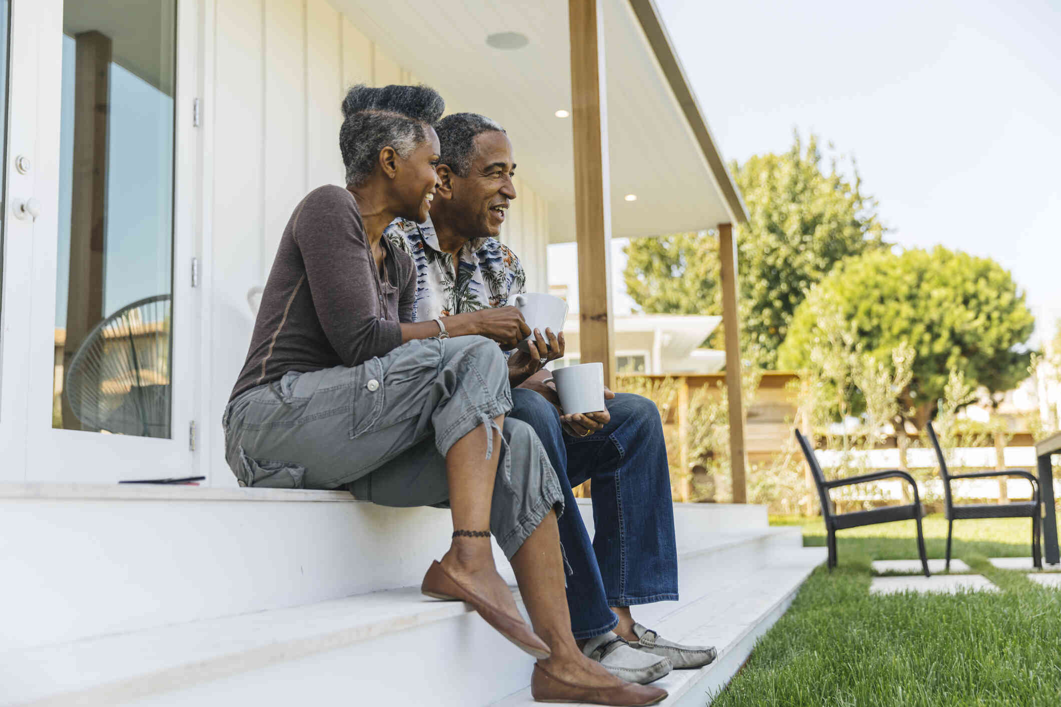 A middle aged male and female couple  sit on their front porch steps with cups of coffee on a sunny day. 