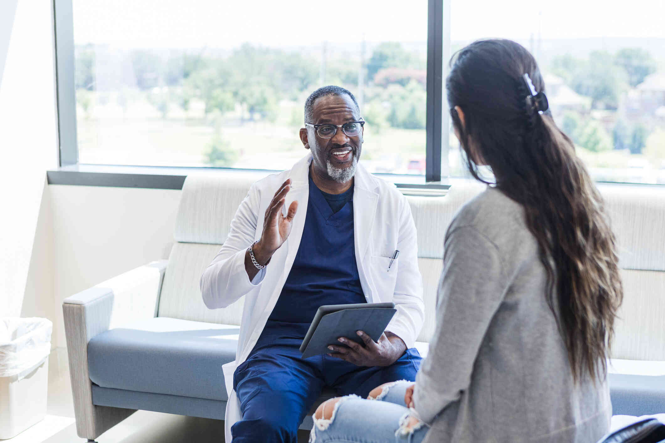 A male doctor in a white lab coat talks while holding aclipboard and sitting across from his female patient.