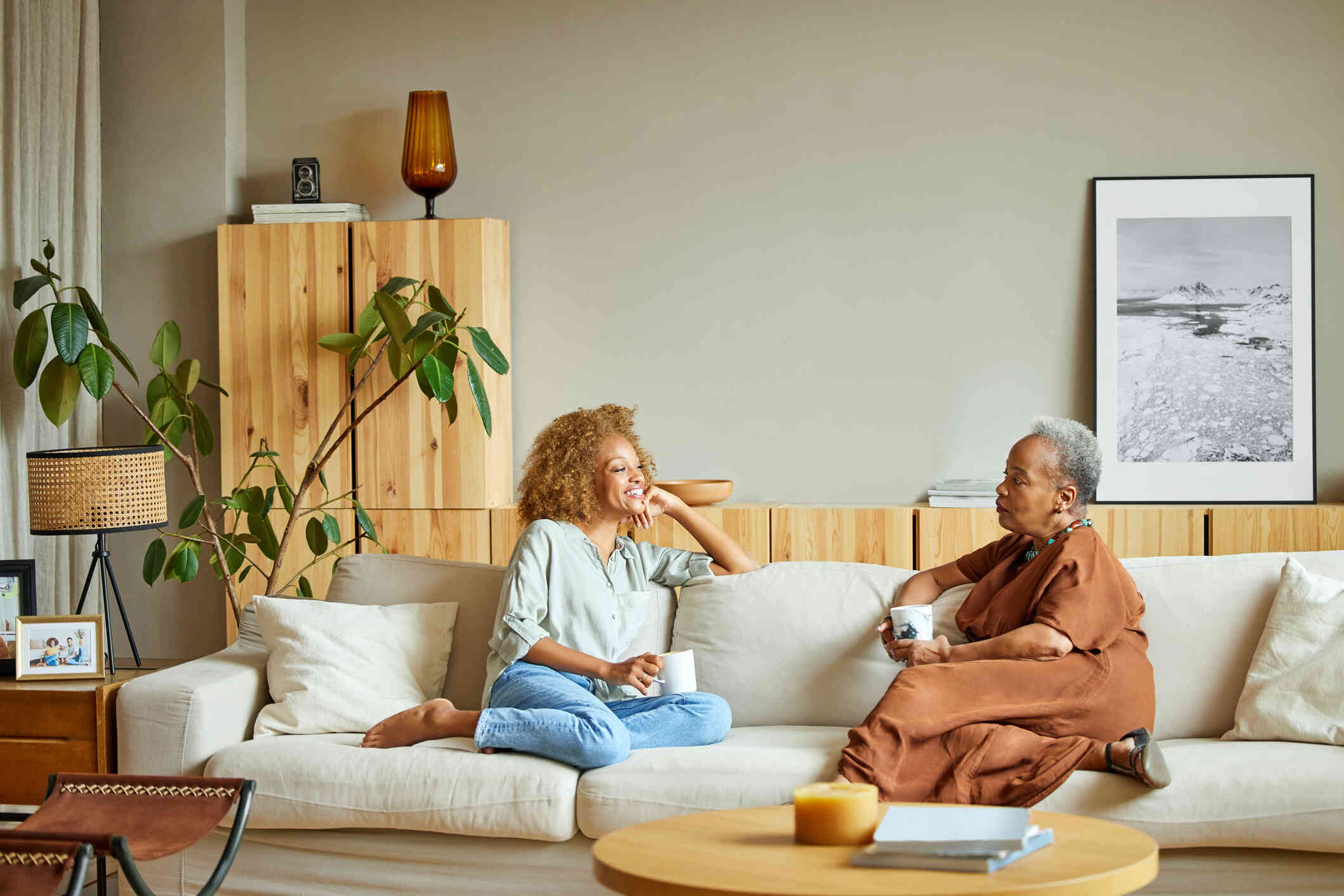 Two female relative sit on a couch facing each other and talk while holding mugs of coffee.